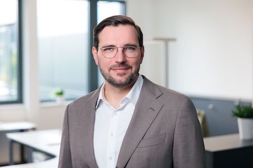 Mann mit braunem Sakko, weißem Hemd und Brille in einem modernen Büro.