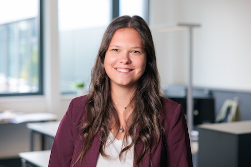 Lächelnde Frau mit langen braunen Haaren trägt einen bordeauxroten Blazer und sitzt in einem hellen Büro.
