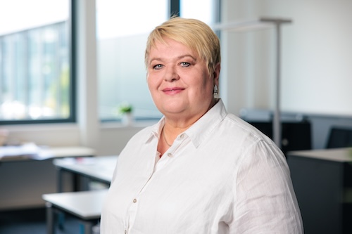 Frau mit kurzem blondem Haar und weißer Bluse lächelt in einem hellen Büro.