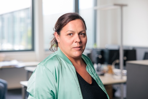 Frau mit grünem Blazer und dunklem Shirt, die in einem Büroumfeld in die Kamera schaut.