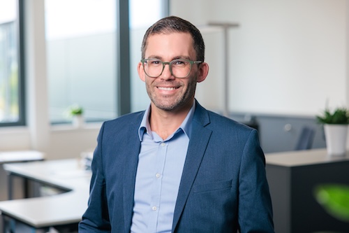 Ein lächelnder Mann mit Brille trägt einen blauen Anzug und ein hellblaues Hemd in einem modernen Büro.