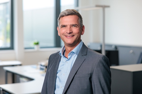 Geschäftsmann mittleren Alters in blauem Hemd und grauem Anzug, lächelnd in einem modernen Büro.
