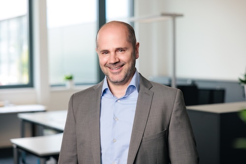 Lächelnder Mann in grauem Anzug und blauem Hemd in einem modernen Büro mit großen Fenstern.