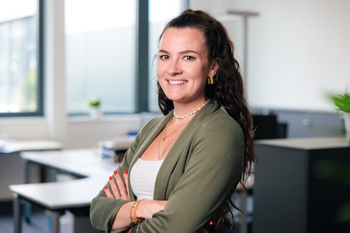 Junge Frau mit langem, lockigem Haar, in einem weißen Top und grüner Jacke, lächelnd mit verschränkten Armen in einem modernen Büro.
