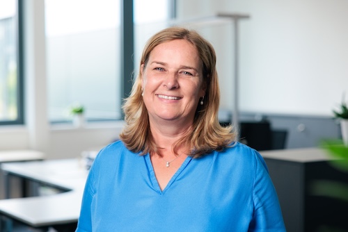 Lächelnde Frau mit blonden Haaren und blauem Oberteil in einem hellen Büro.