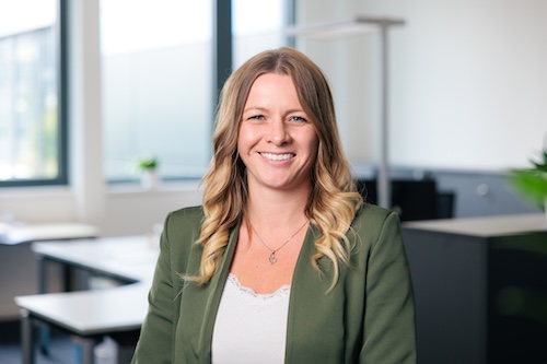 Lächelnde Frau mit langen, welligen, blonden Haaren in einem grünen Blazer in einem modernen Büro.