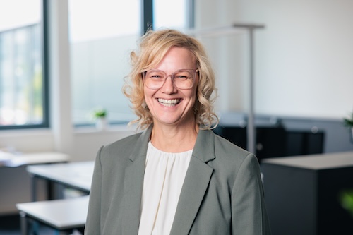 Lächelnde blonde Frau mit Brille und grauem Blazer in einem modernen Büro.