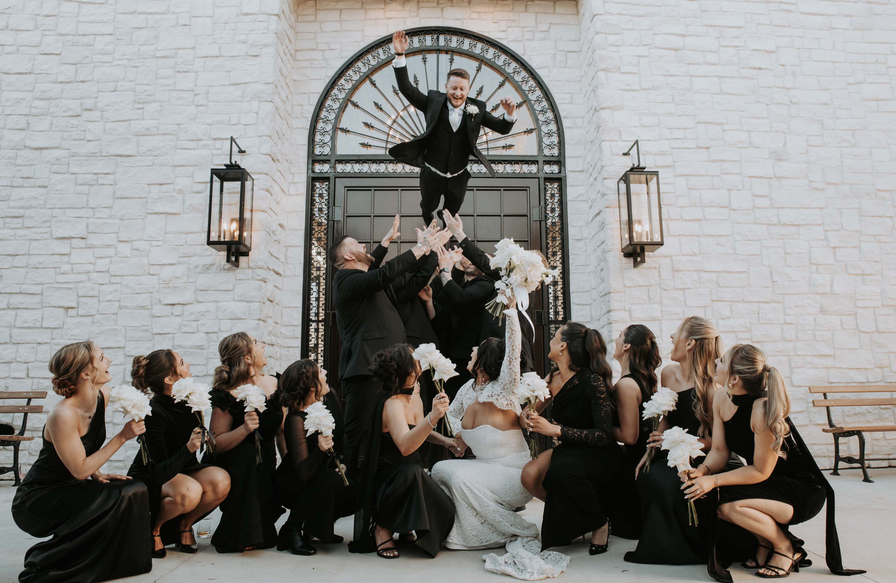 Groom jumps through archway into cheering bridal party at a wedding venue.