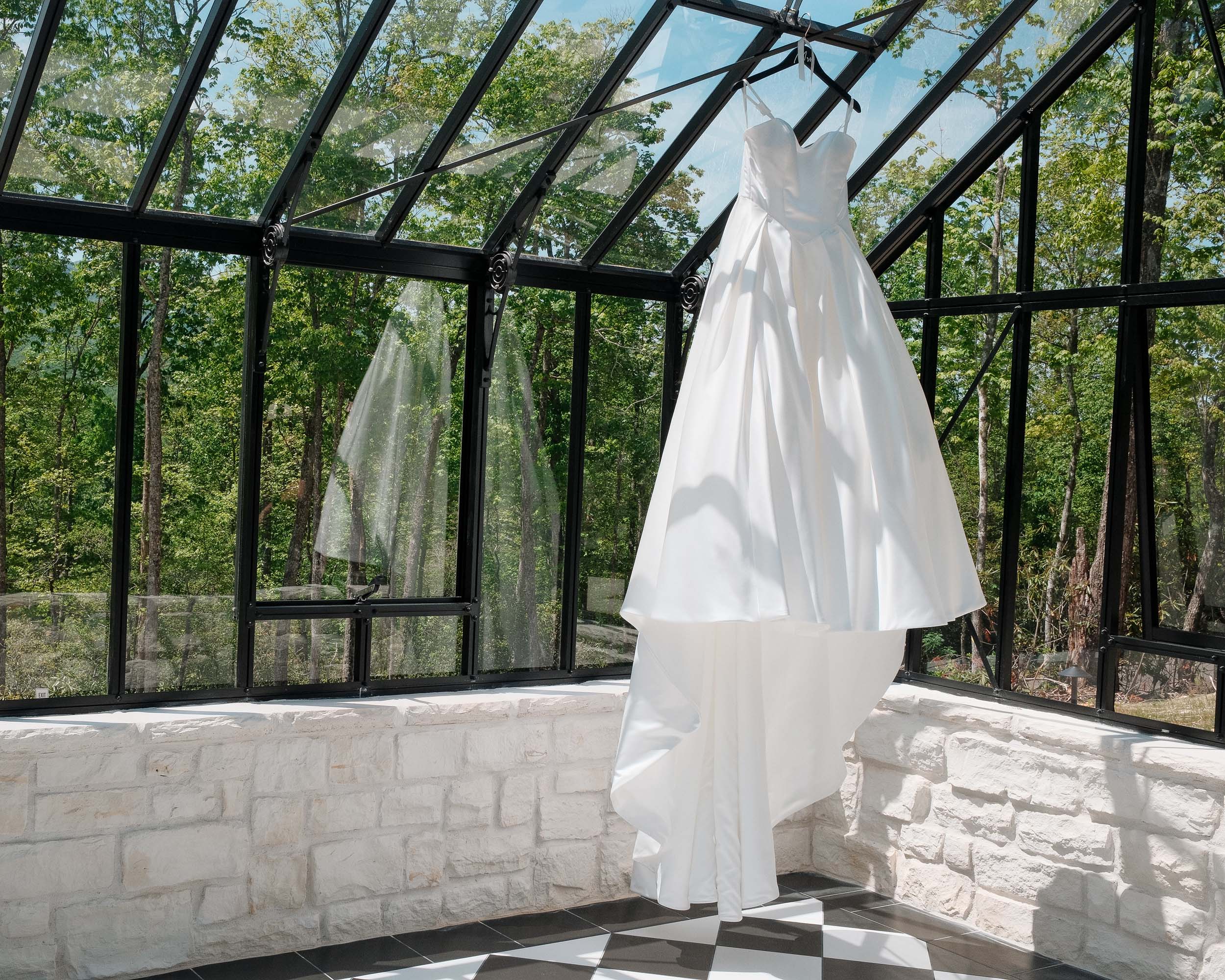White wedding dress hanging in a glass-enclosed venue overlooking woods.