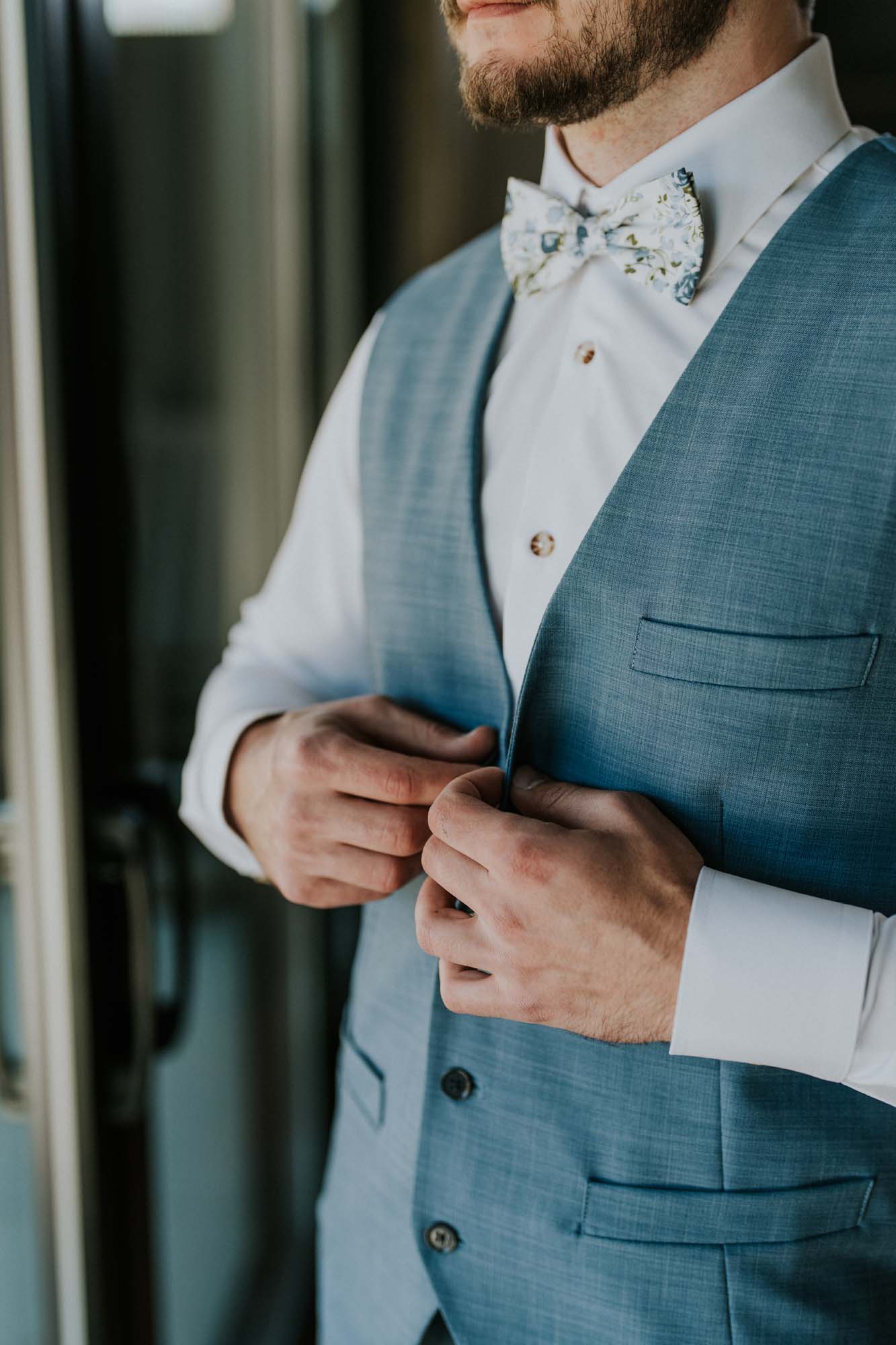 Groom adjusting his blue suit jacket with a patterned bow tie.