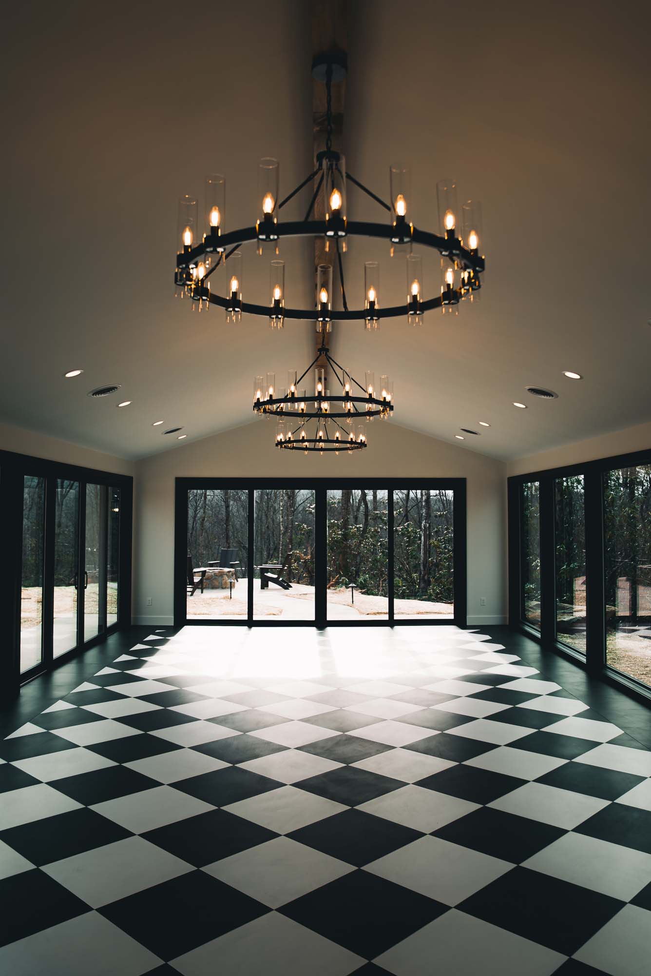 Grand hall with black and white checkered floor, chandelier, and large windows overlooking woods.