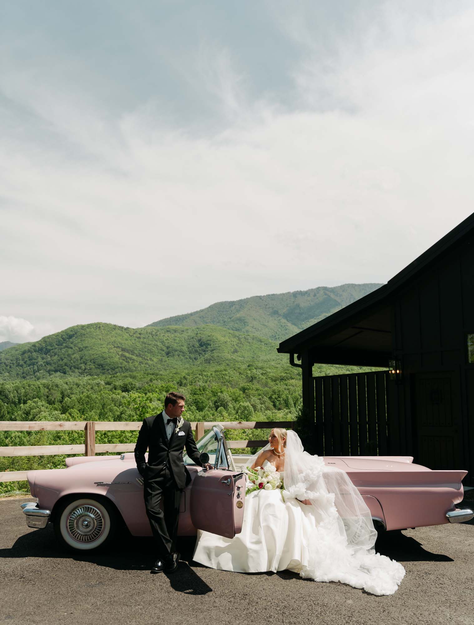 Newlyweds pose beside a vintage pink car with mountains in the background at Trailhead Venues.