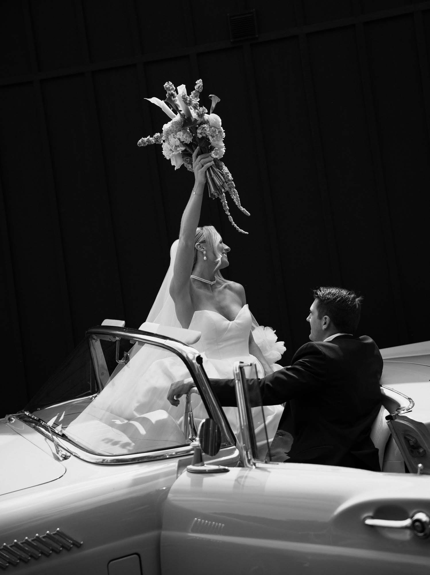 Bride in white dress raises bouquet while groom watches, suggesting a wedding venue moment.