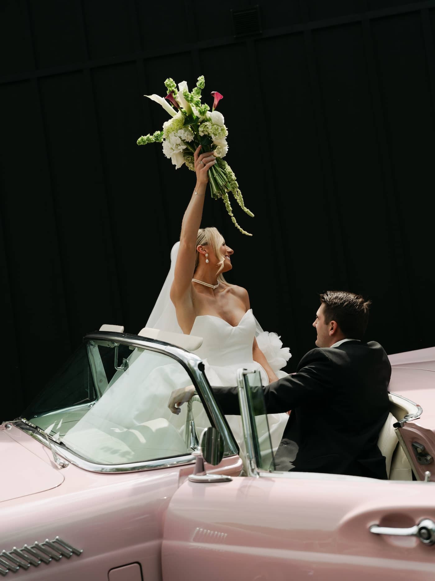 Bride in white dress holds bouquet, looking at groom by vintage car.