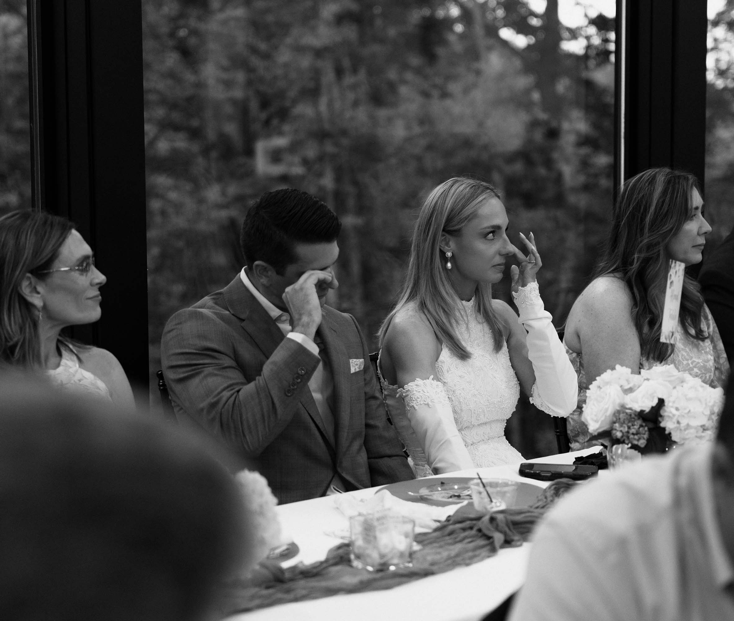 Three women and a man seated indoors by a large window overlooking trees.