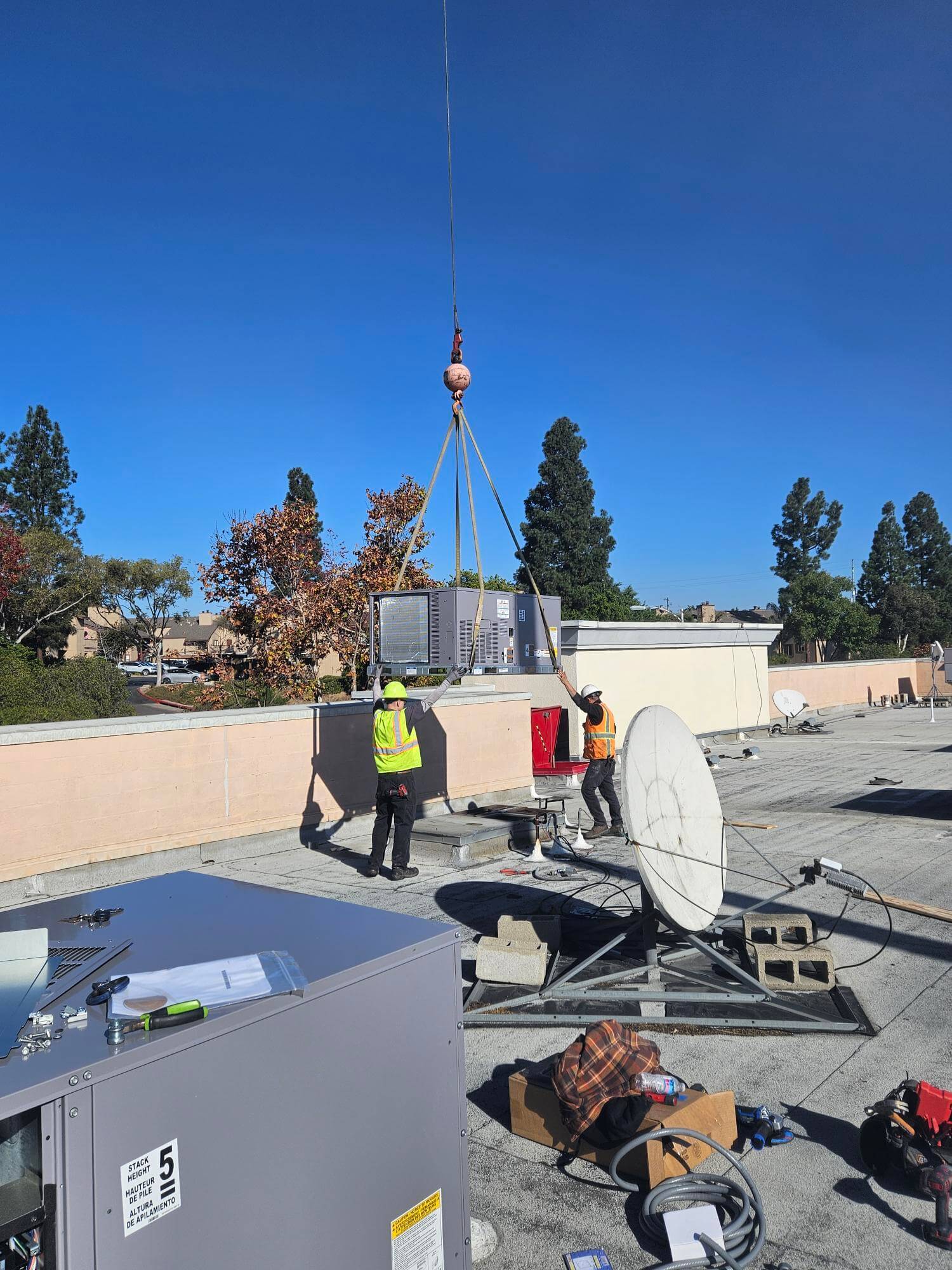 Two workers guide a crane lowering an air conditioning unit onto a building roof.