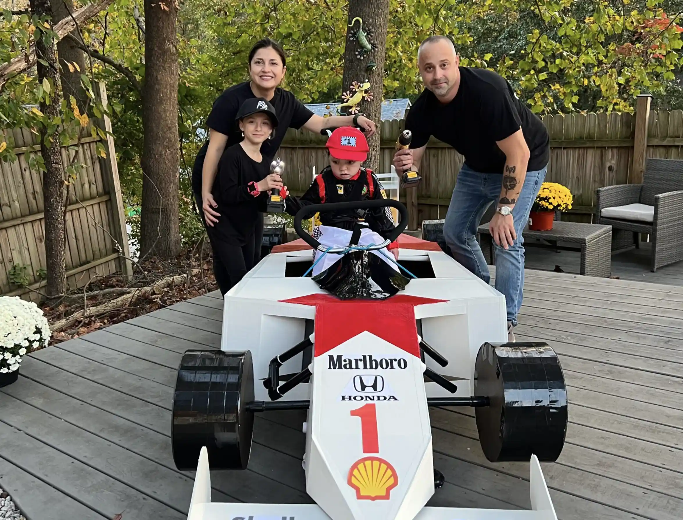 A child dressed as a race car driver sitting in a white and red cardboard Formula 1 car with Marlboro and Honda logos, flanked by a smiling woman and man holding toy drills and another child in black.
