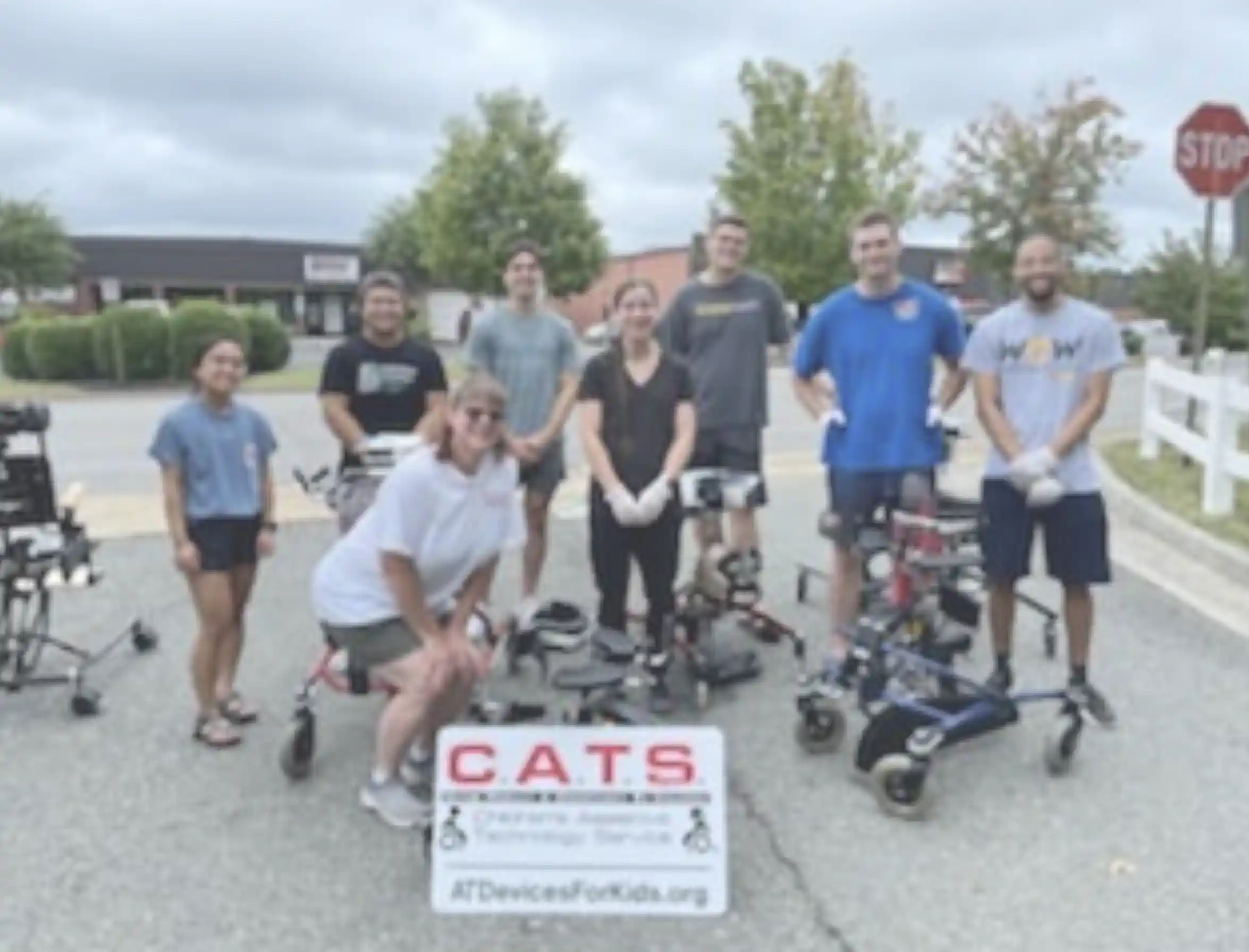 Group of eight people posing outdoors with assistive walking devices and a sign reading CATS Assistive Technology Services.