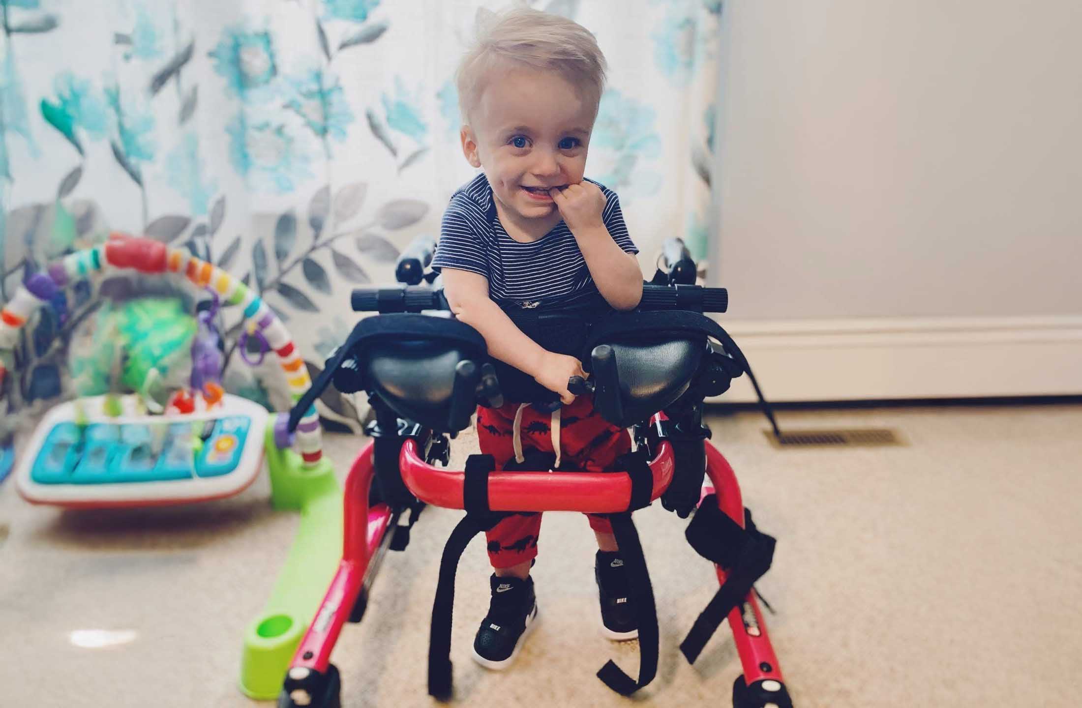 Smiling toddler in striped shirt using a red mobility walker in a living room.