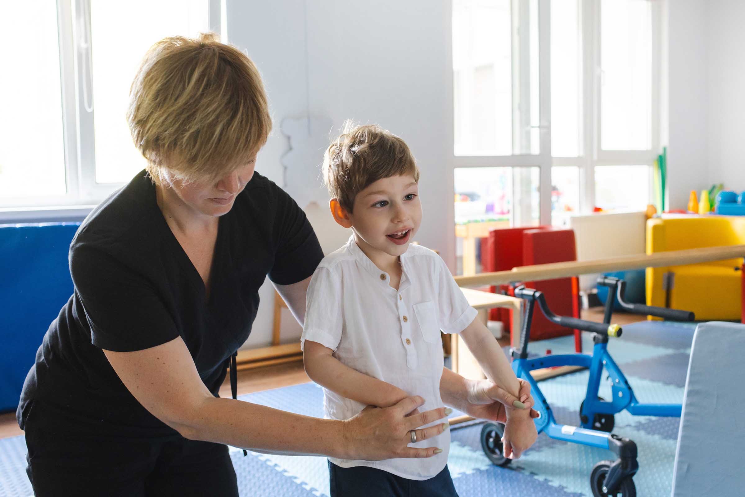 Therapist assisting a smiling young boy with walking exercises in a brightly lit therapy room.
