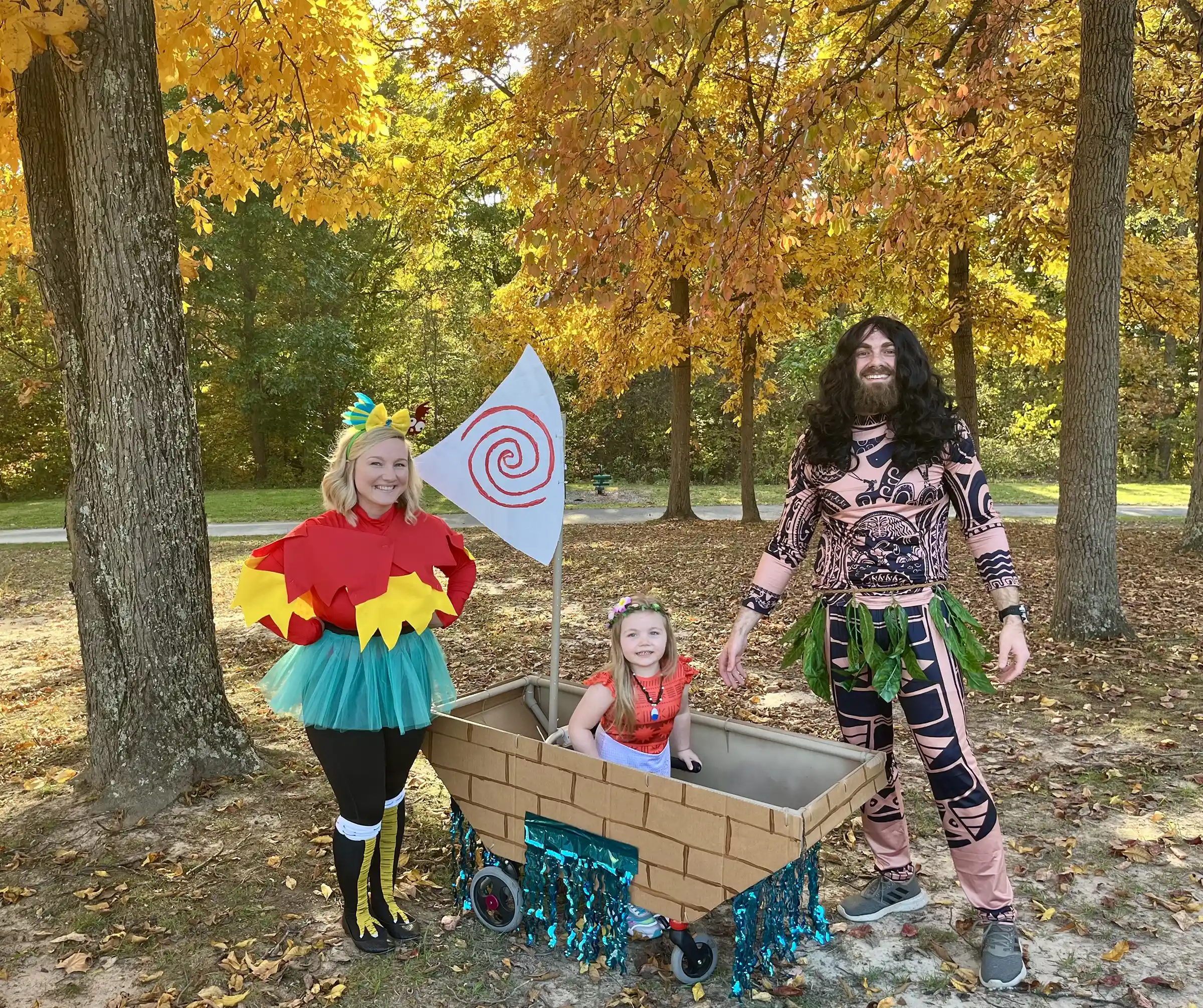Three people dressed in Moana-themed costumes, including a child in a boat decorated with a sail featuring a red spiral symbol, standing outdoors under fall foliage.