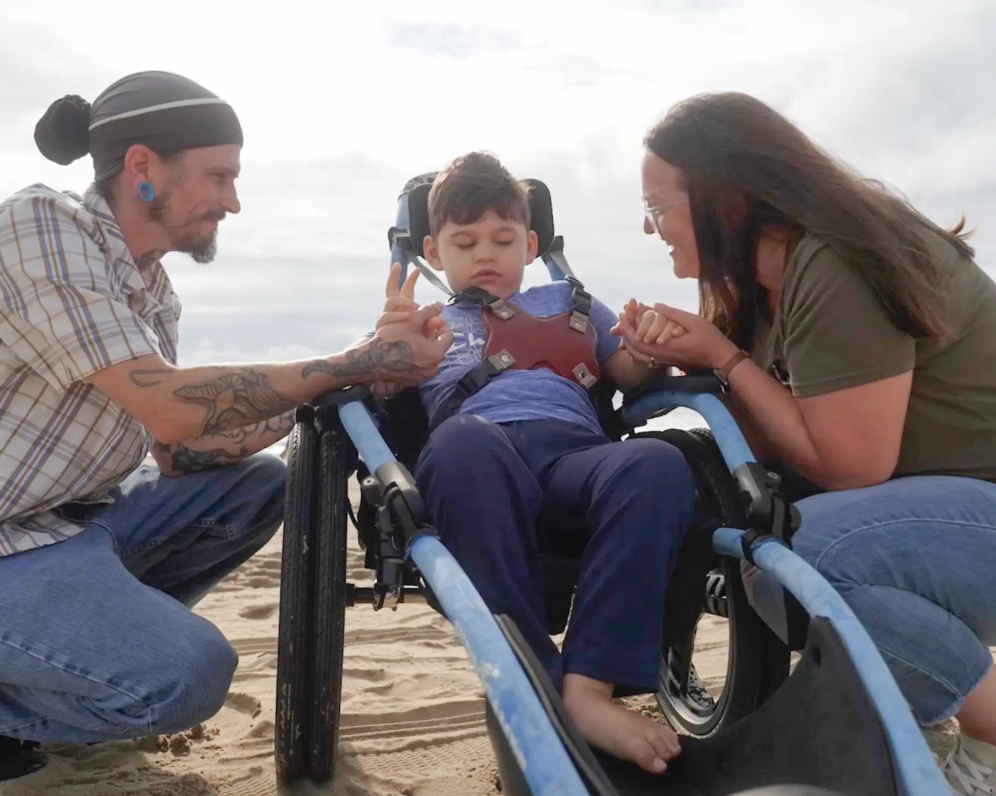 A child in a specialized wheelchair on sand being supported and held by a smiling man and woman on either side.