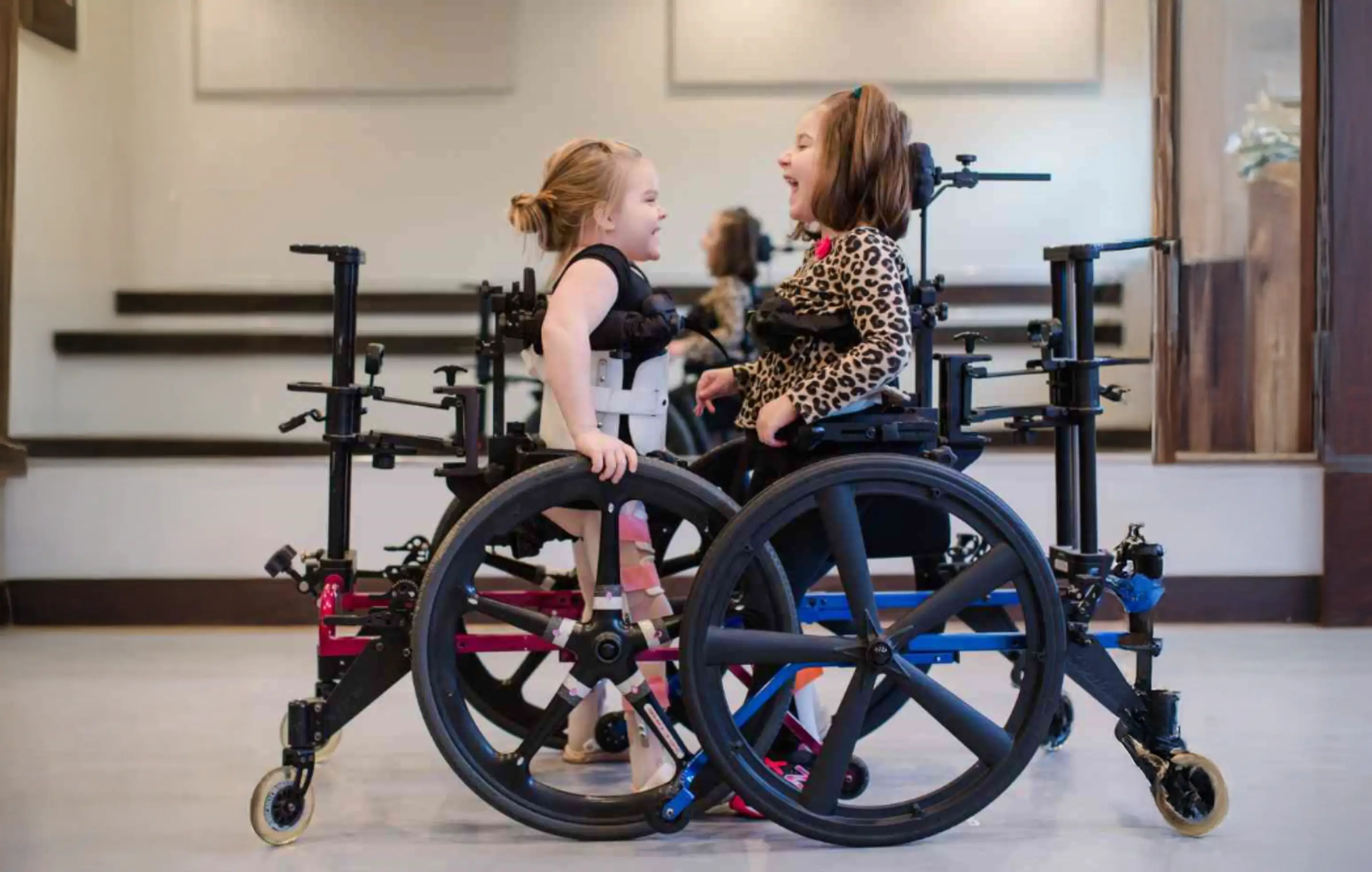 Two young girls in wheelchairs facing each other and smiling in a room with mirrors.
