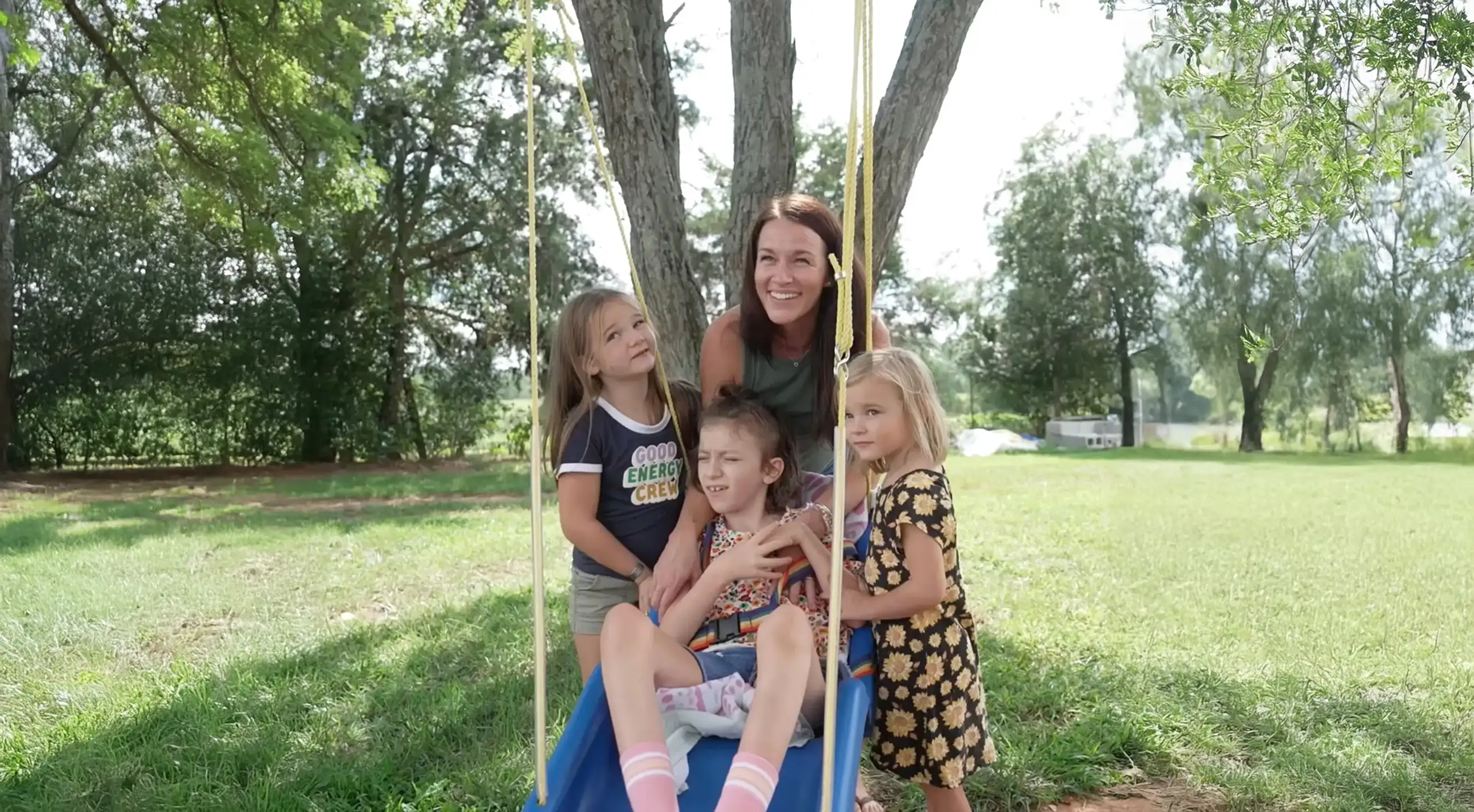 A woman smiles behind three young girls, one sitting on a swing and two standing beside her in a sunny park.