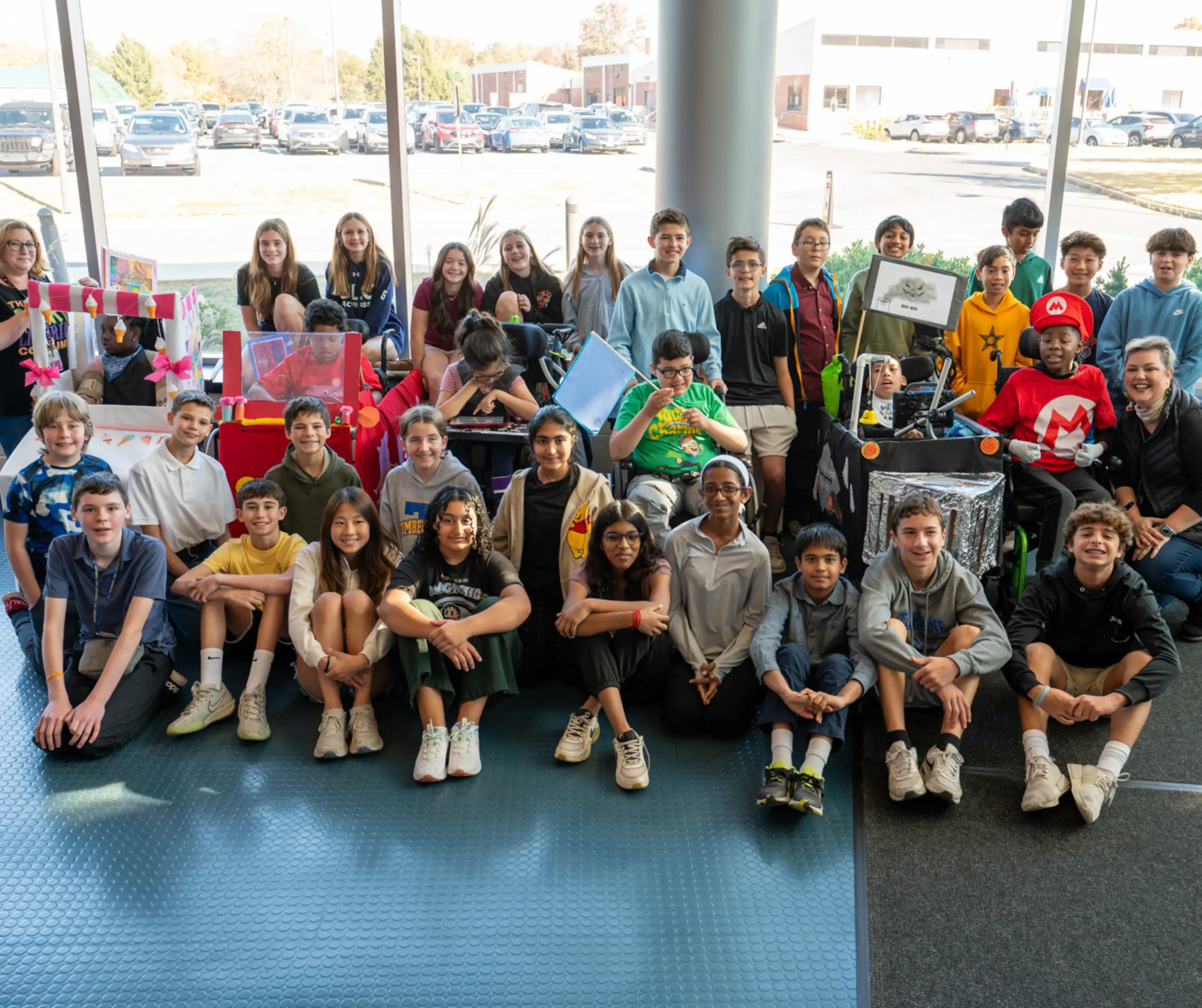 Large group of diverse children and teenagers posing indoors near large windows with decorated wheelchairs and costumes.