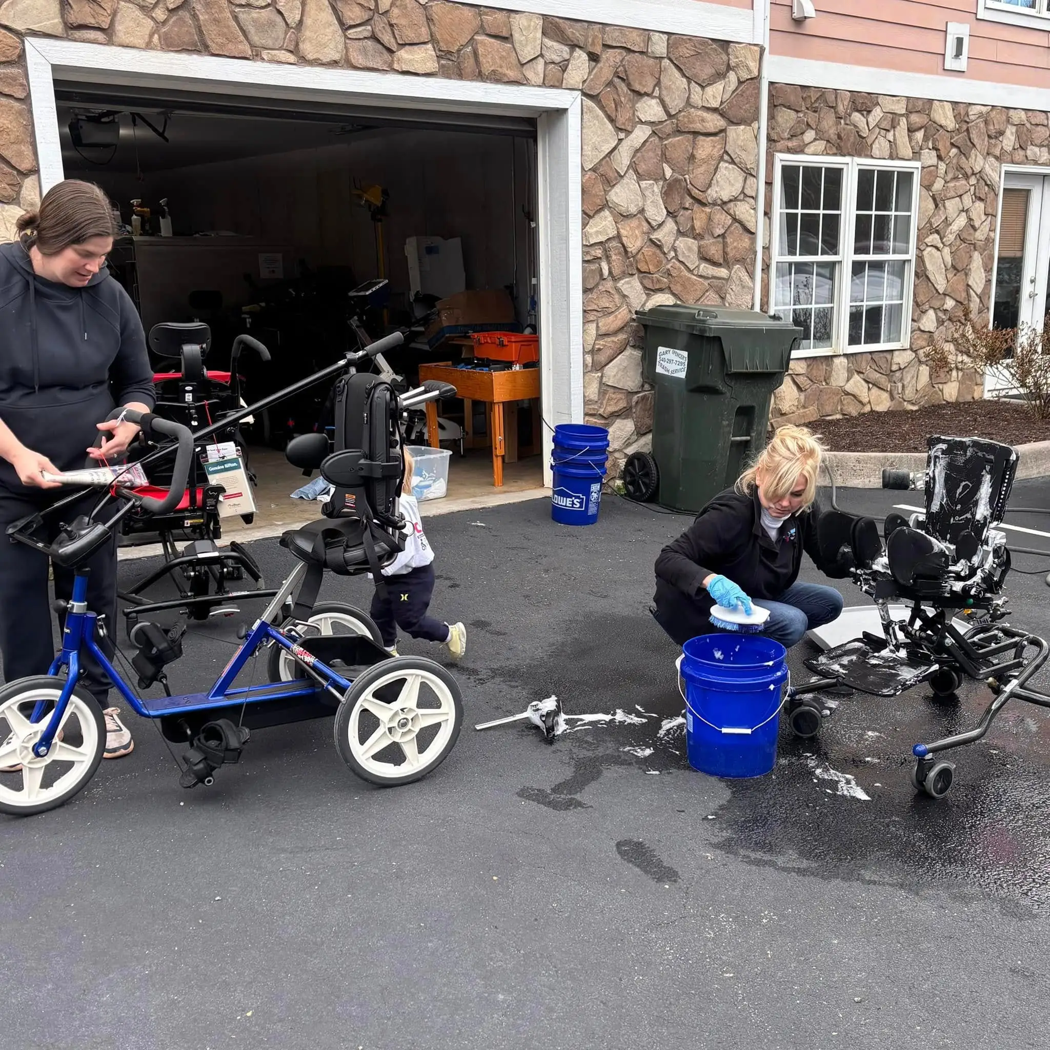 Two women cleaning adaptive bicycles outside a stone building garage, one scrubbing a blue bucket and the other drying a bike part.