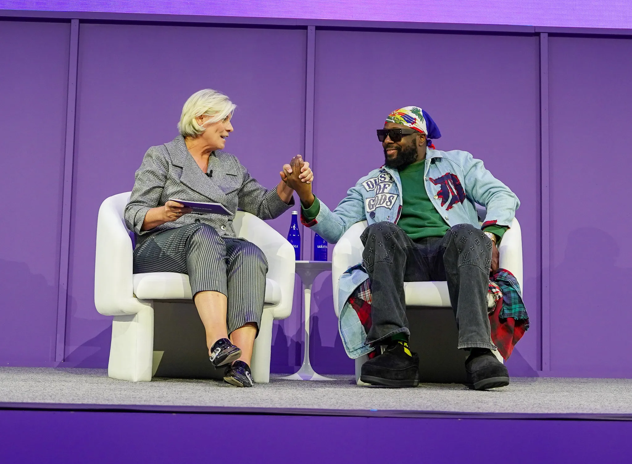 Two people sitting on white chairs on stage against a purple background, holding hands and smiling.