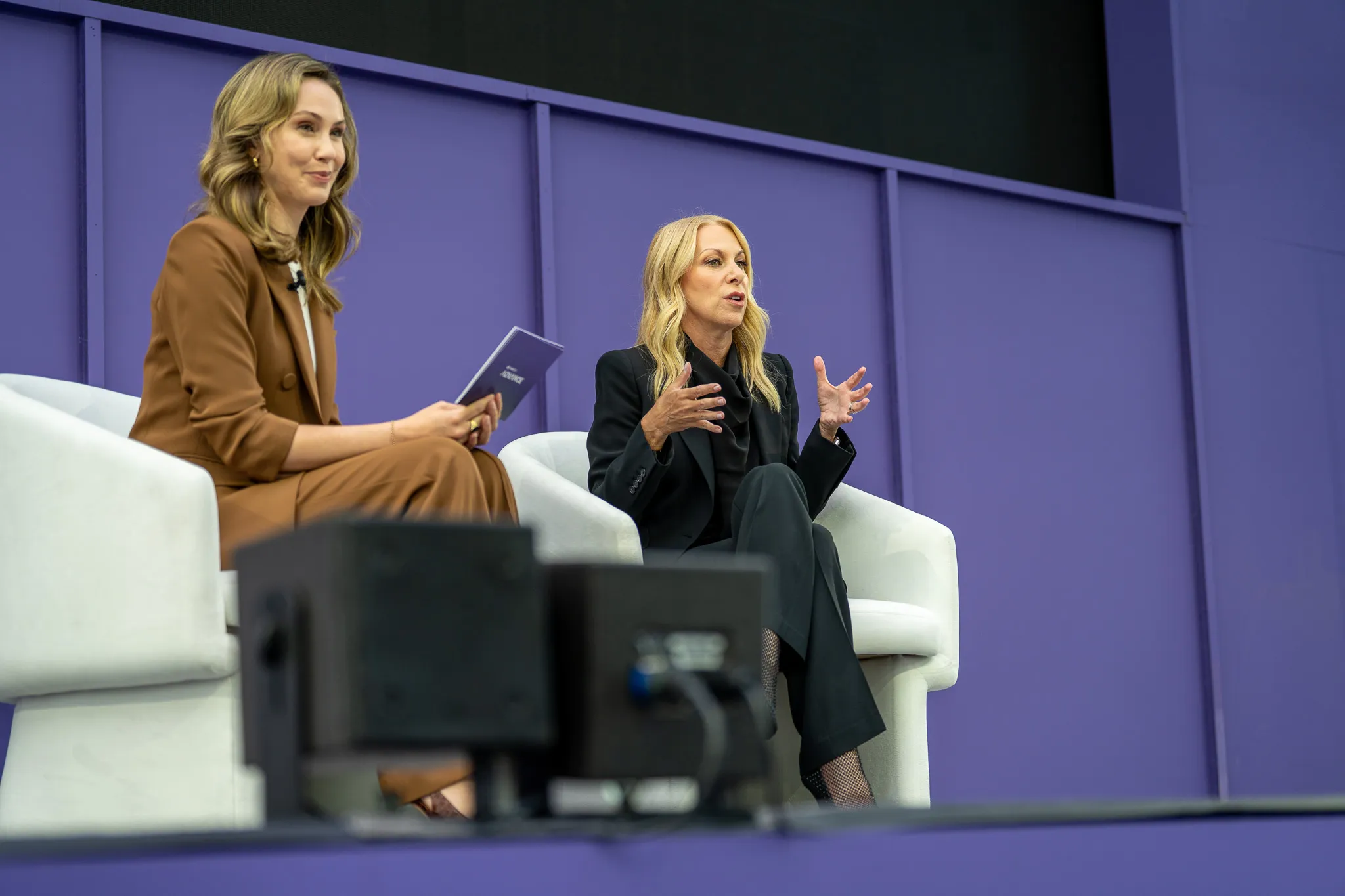 Two women seated on white chairs on stage, one in a brown suit holding a notebook and the other in black speaking with expressive hand gestures.