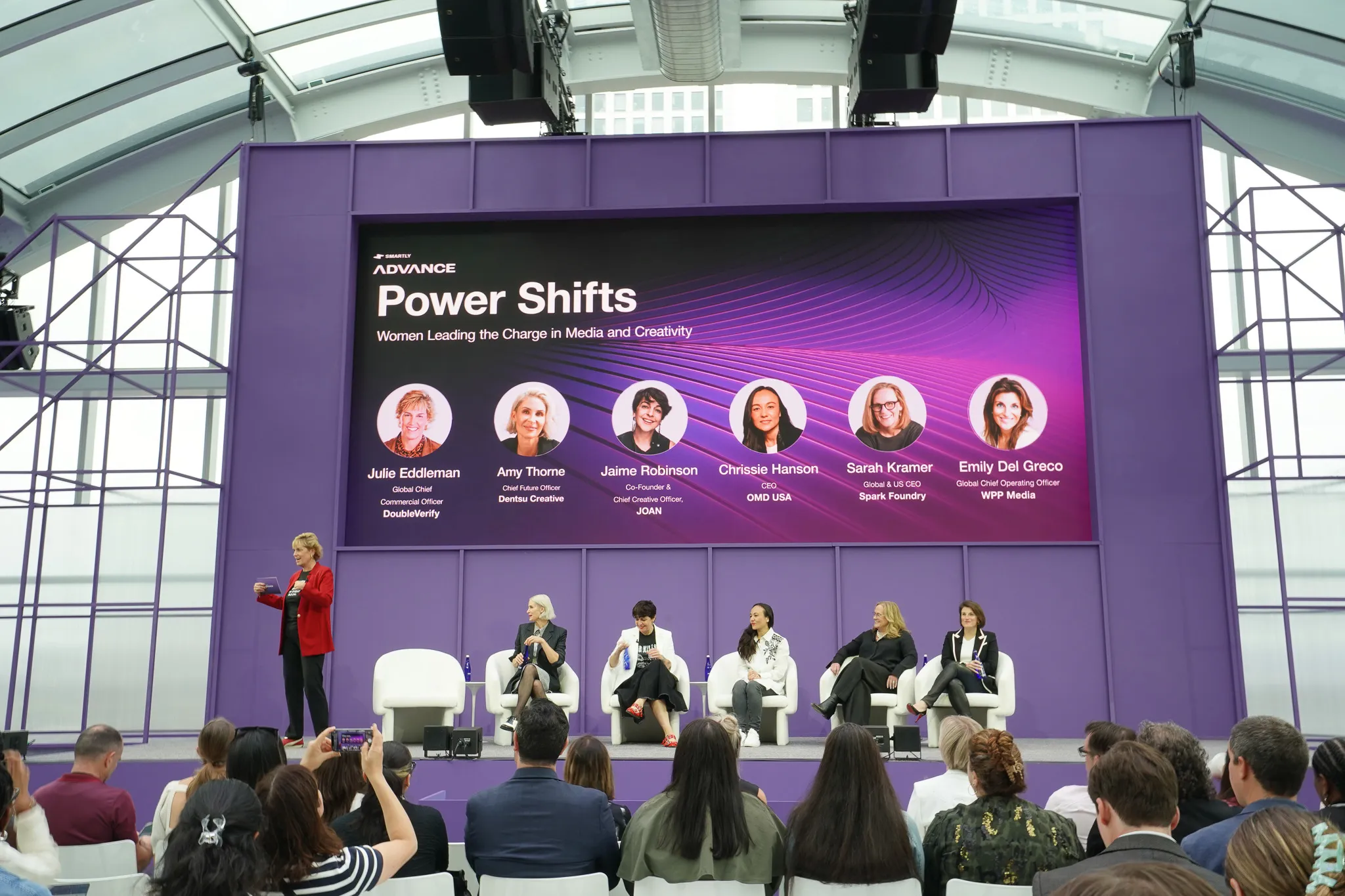Audience seated in rows inside a bright indoor venue with slanted glass ceiling and purple banners.