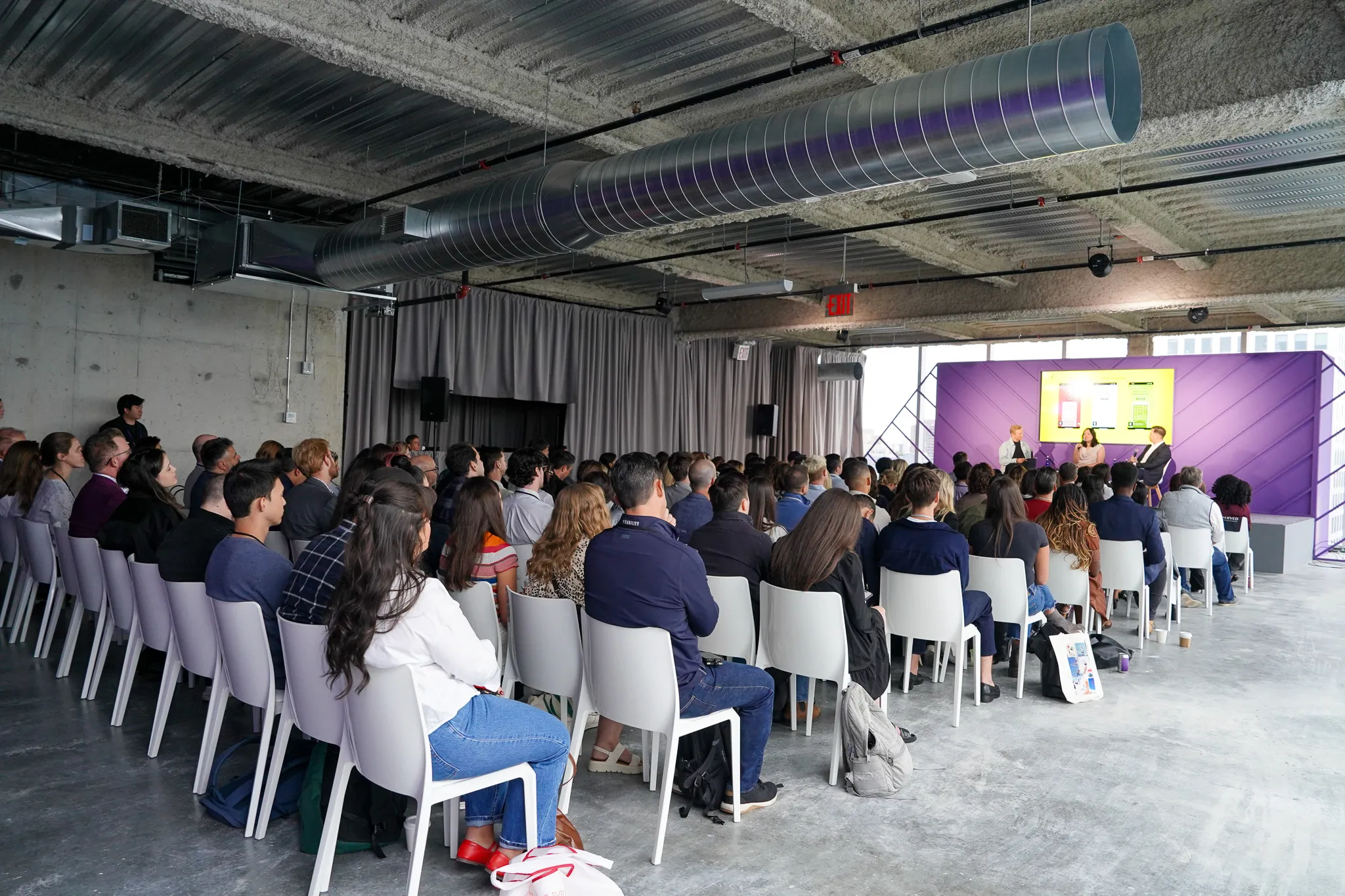 Audience seated indoors facing a stage with speakers in front of a purple backdrop and screen.