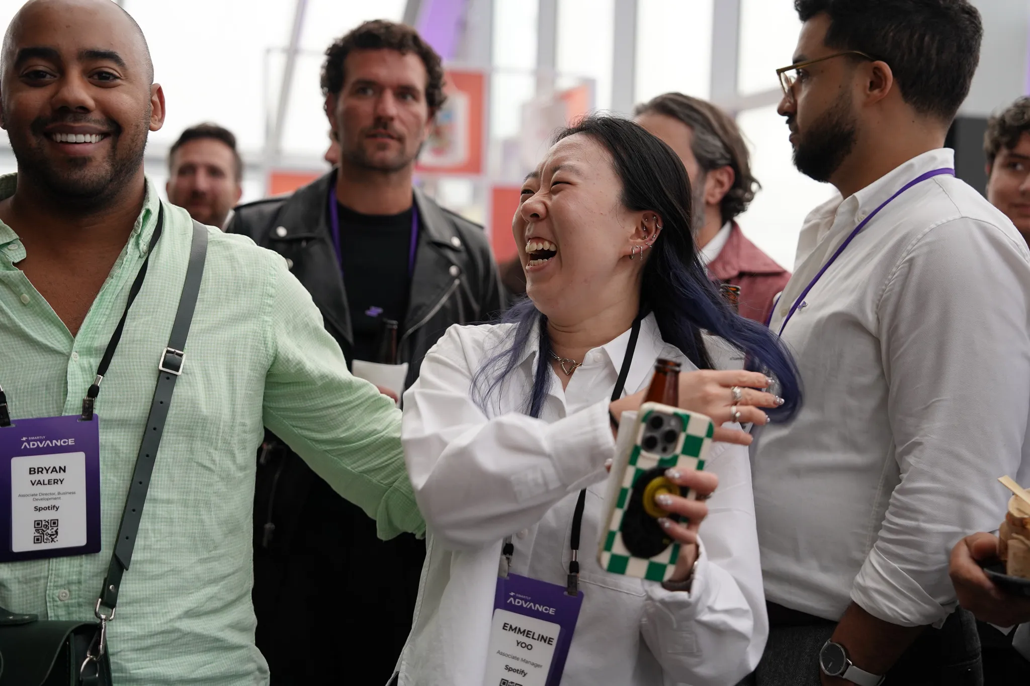 Group of event attendees laughing and socializing with name badges visible, one woman holds a phone and a drink.