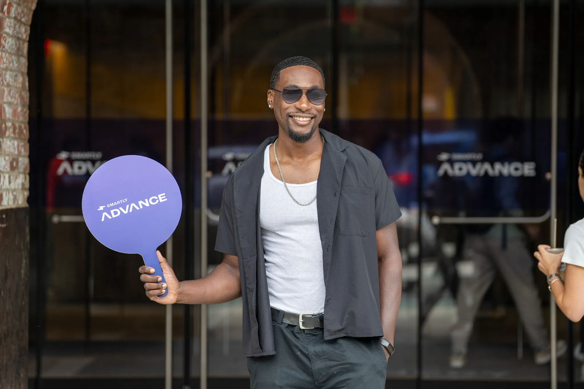 Man smiling, holding a purple Smartly Advance sign paddle at an event entrance with multiple Smartly Advance logos behind him.