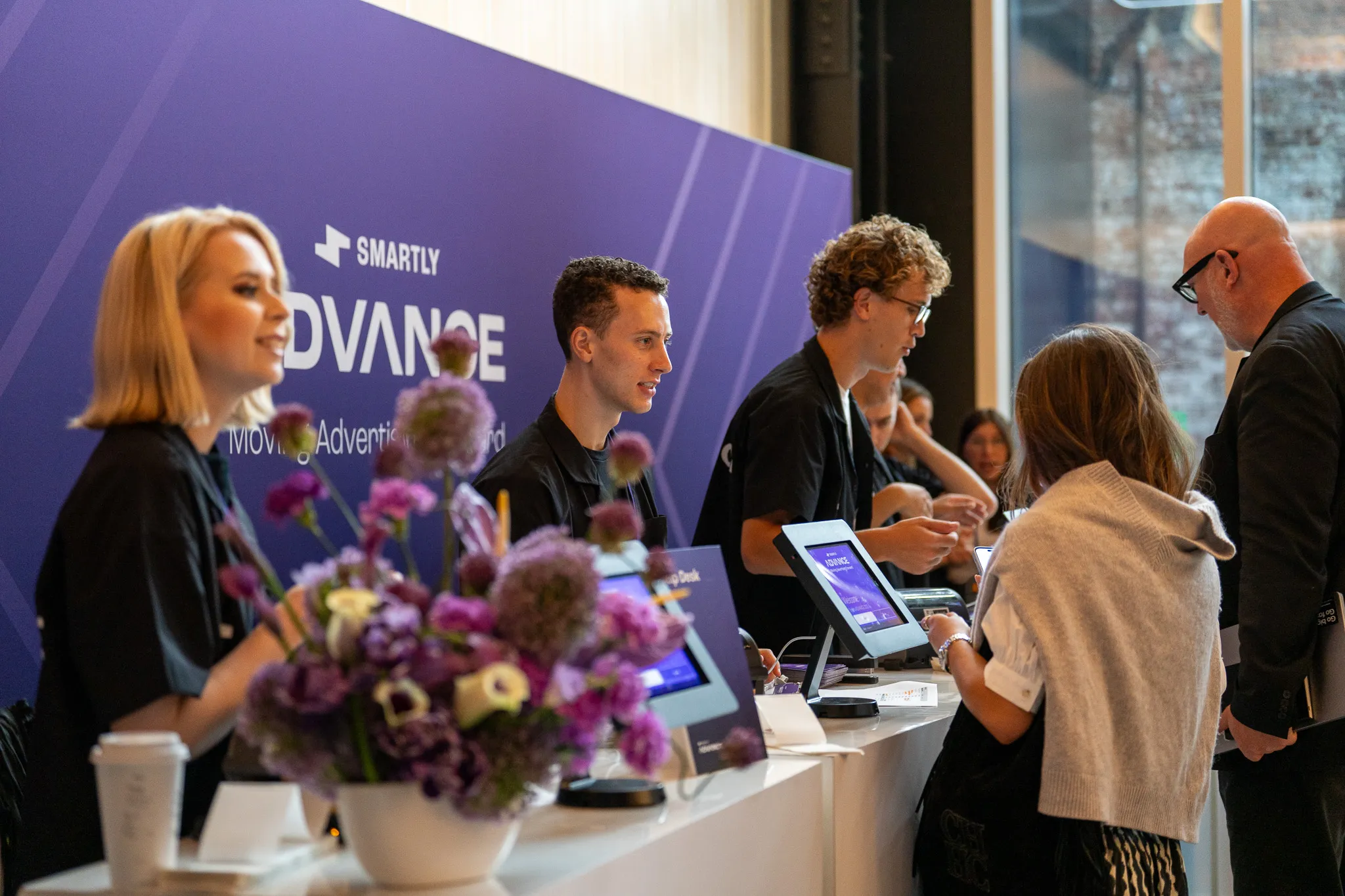 Busy event registration desk with staff and attendees interacting, with Smartly Advance signage in the background and floral decorations.