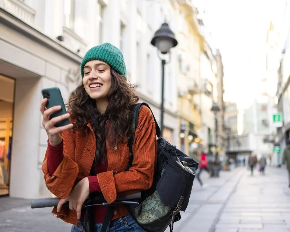 Smiling young woman in green beanie and orange jacket using phone while leaning on scooter in city street.