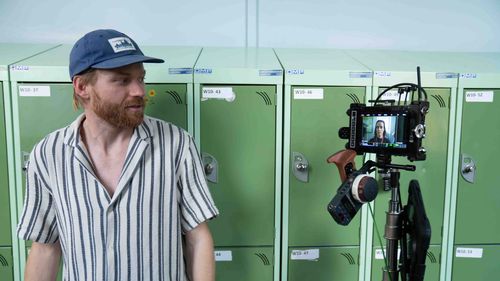 Man in striped shirt and blue cap standing near green lockers with a video camera recording a woman visible on the camera screen.