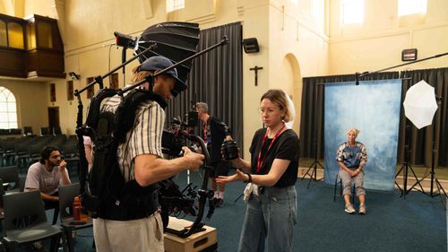 Photography studio setup inside a hall with a woman sitting in front of a blue backdrop and a man operating a camera rig while a woman adjusts camera equipment nearby.