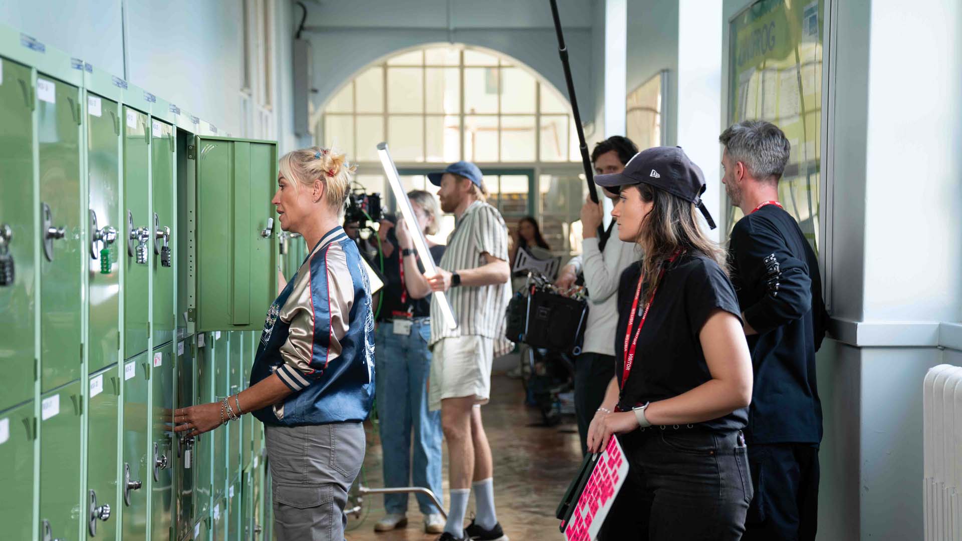 Film crew shooting a scene where a woman opens a green school locker in a hallway.