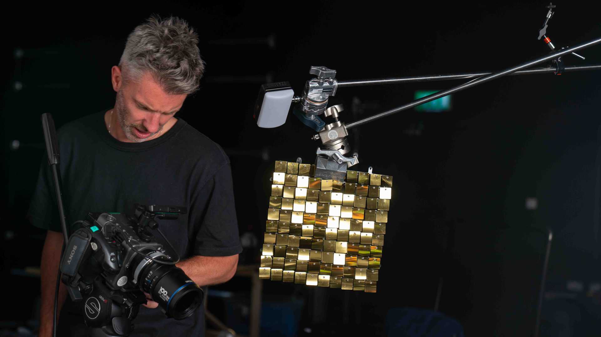 Man operating a professional Sony video camera filming a reflective gold square panel suspended by metal arms in a dark studio.