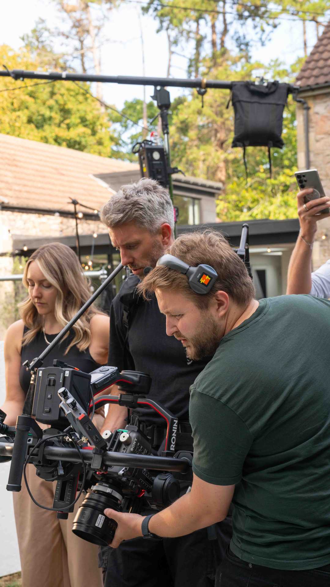 Three people operating professional camera equipment outdoors with trees and a building in the background.