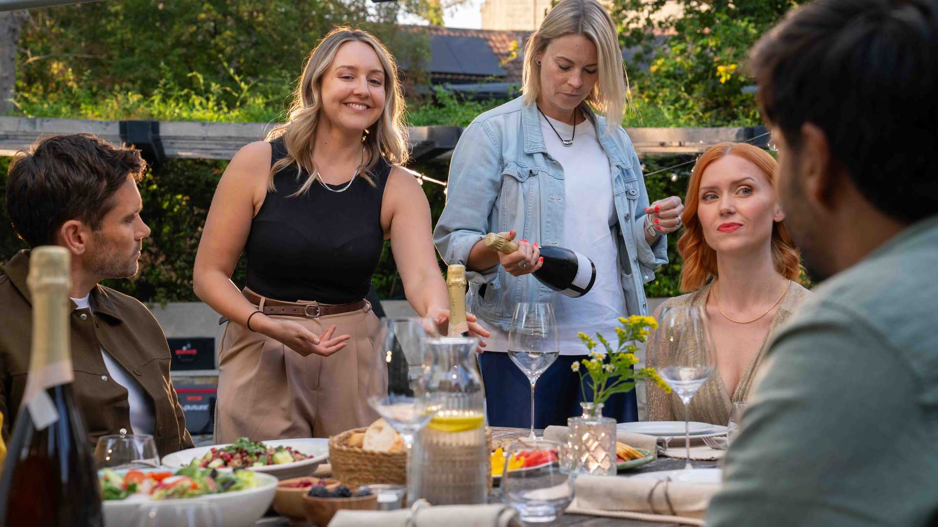 Group of five adults enjoying an outdoor meal with wine, one woman smiling and reaching for a bottle while another woman pours wine into a glass.