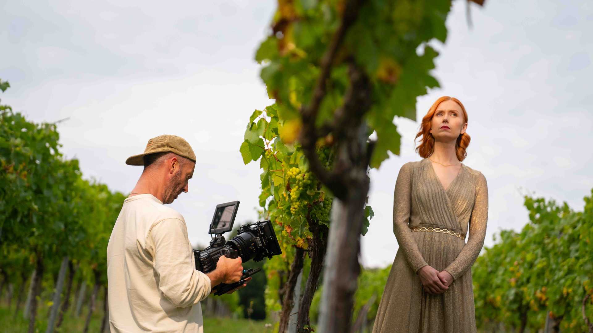 Man in a cap filming a woman in a long gold dress standing among rows of grapevines.