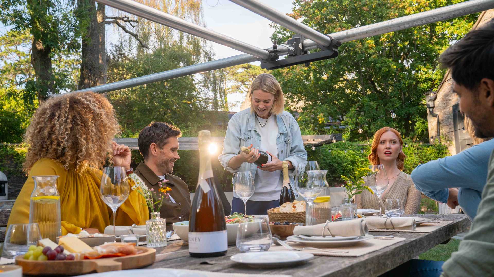 Group of five friends gathered outdoors around a wooden table with food and drinks, one woman opening a bottle of champagne.