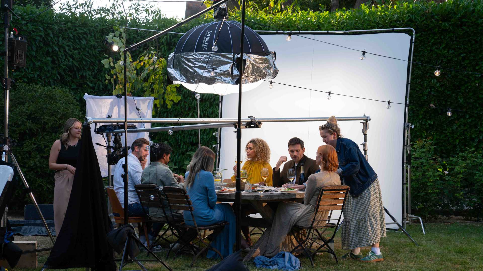 Film crew setting up a dinner scene with actors seated around a table outdoors, surrounded by lighting and camera equipment.
