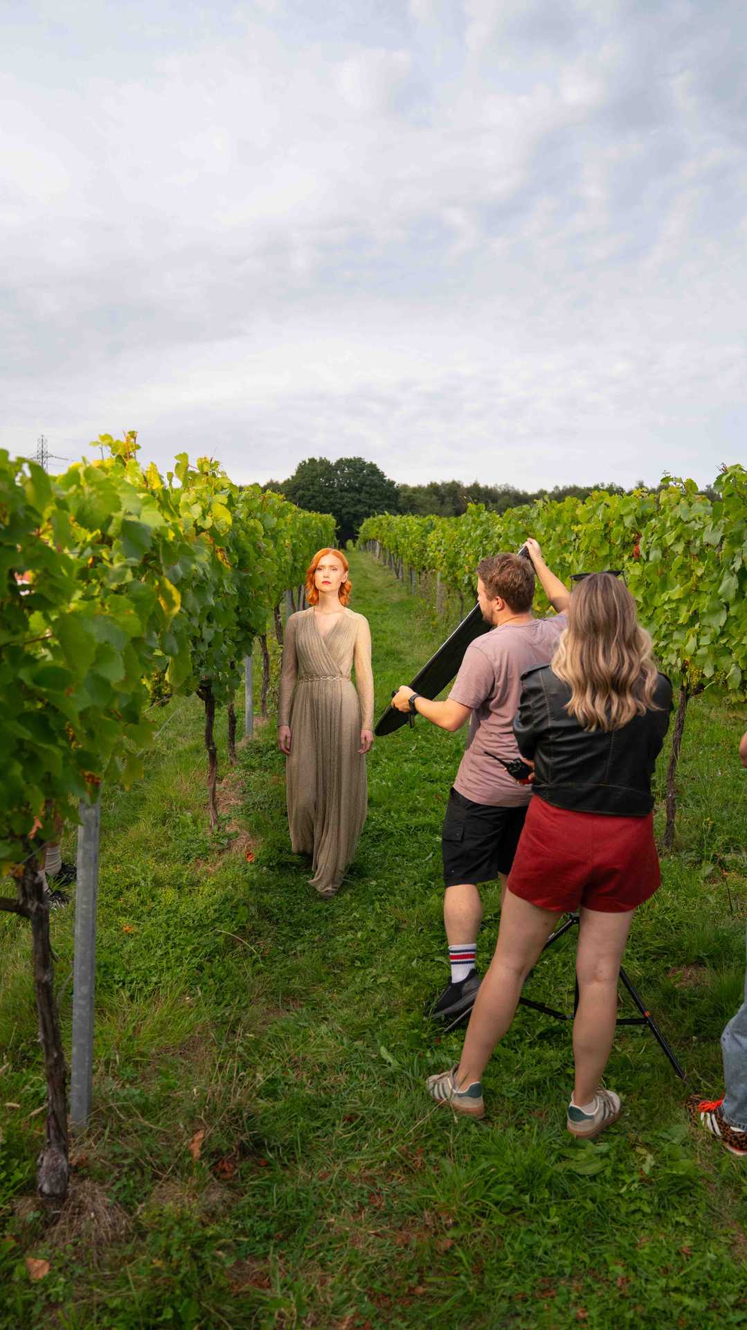 A red-haired woman in a long beige dress posing between green vineyard rows while two people adjust lighting and camera equipment.