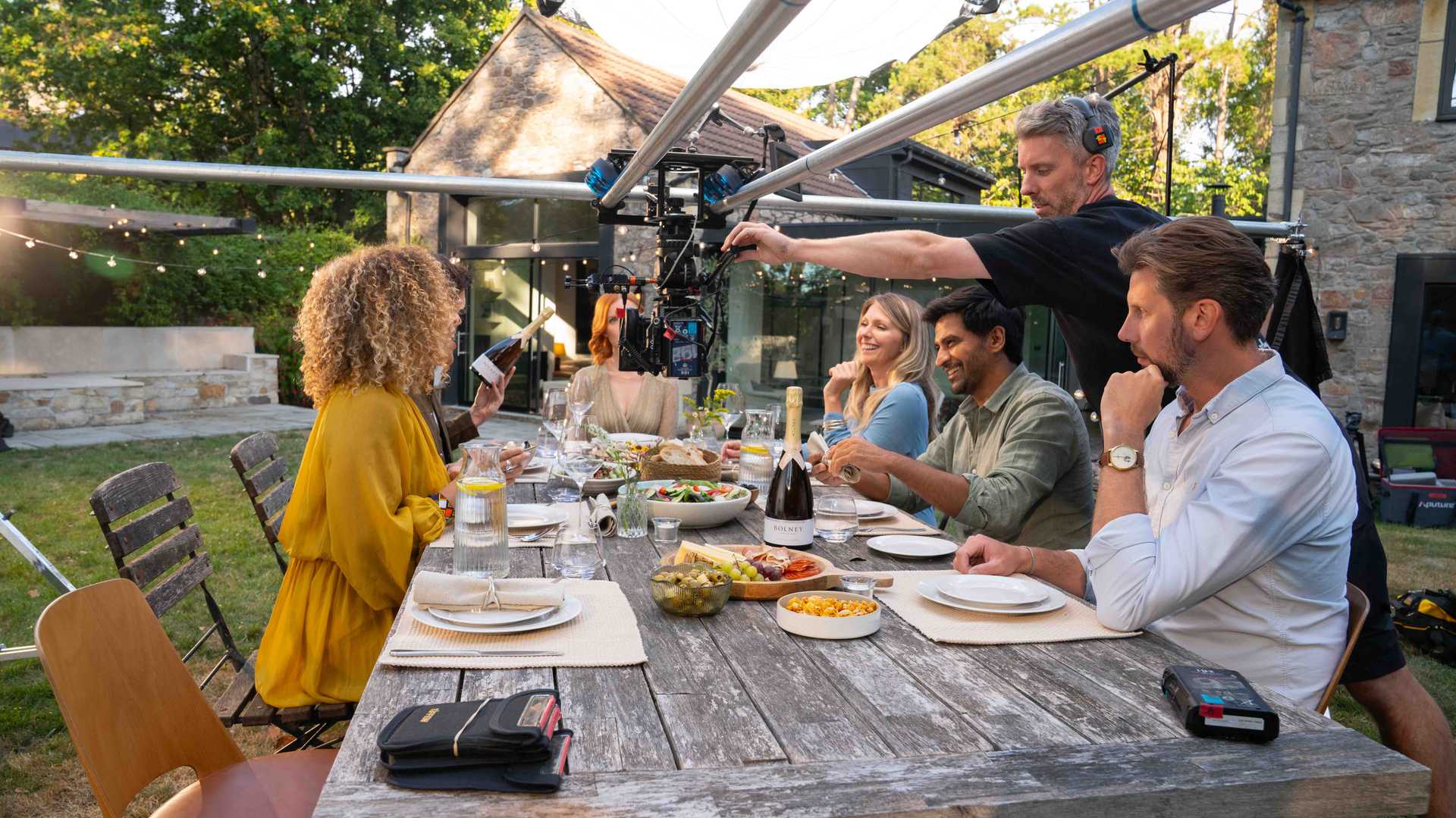 Group of people sitting at an outdoor table with food and drinks, being filmed by a cameraman using a rig.