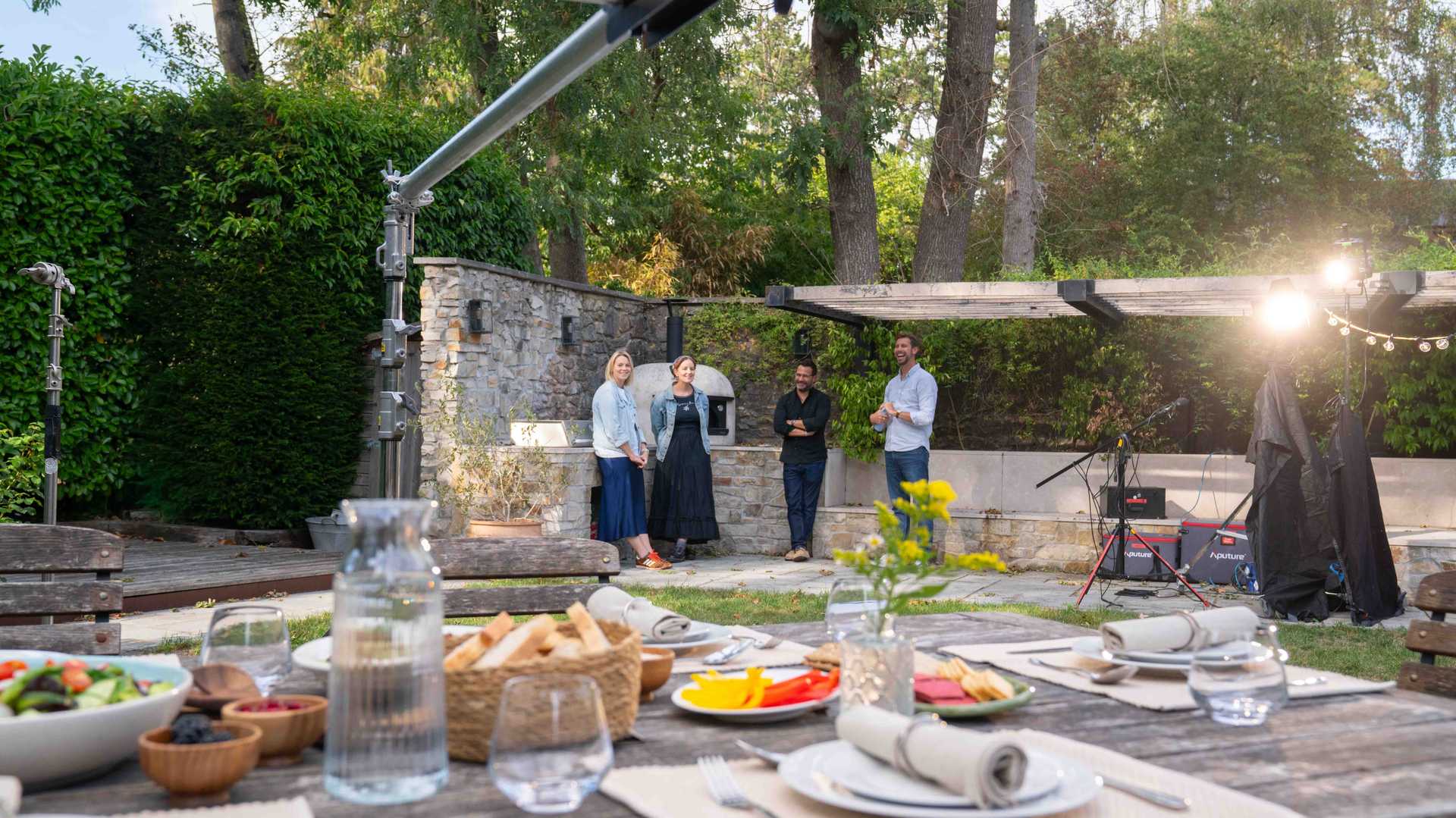 Four people standing and talking outdoors near a stone oven in a garden with a wooden table set with food and drinks in the foreground.